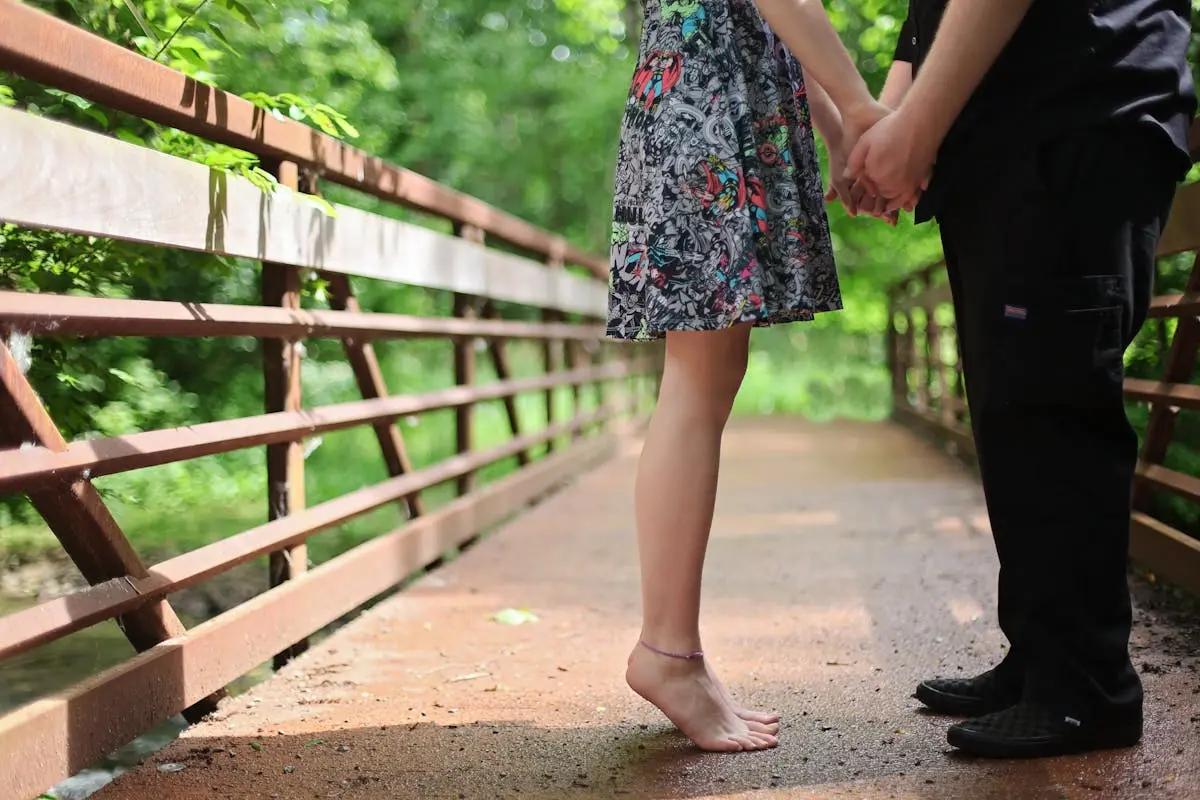 Une femme sur la pointe des pieds tenant les mains d’un homme sur un pont en bois entouré de nature, symbolisant l’intimité et la connexion dans le couple.